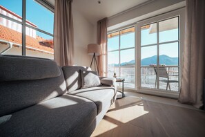 A modern hotel room with a sofa, a lamp, a small table, and a view of a balcony with outdoor seating.