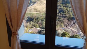 Uma janela com cortinas, vista de uma paisagem montanhosa e um edifício com telhado de azulejos.