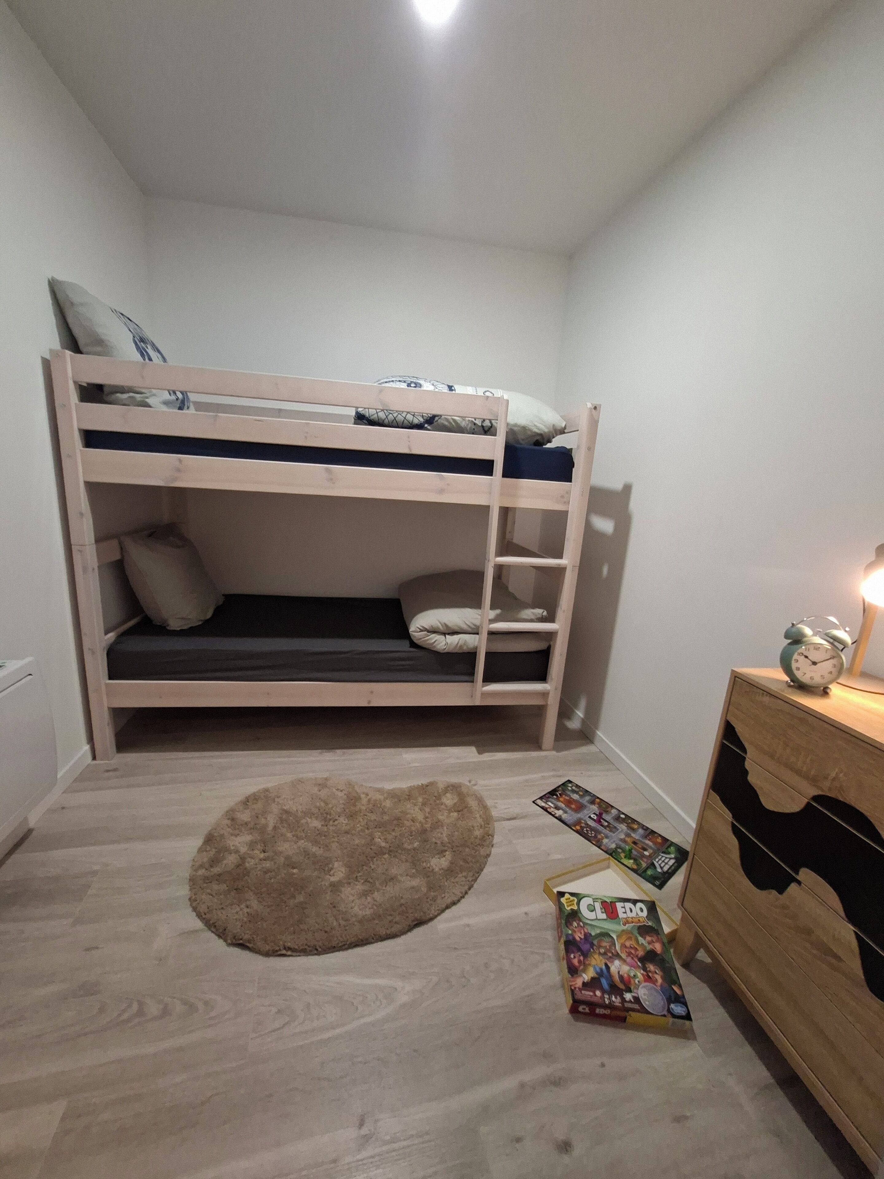 A bunk bed with a wooden frame, a small rug, a dresser, and a clock on the dresser.