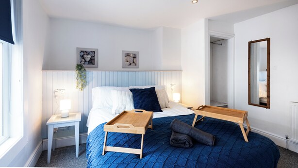 A bedroom with a bed, bedside tables, a bedside lamp, a hanging plant, a mirror, and a door leading to another room. - Guestz at The Wellington (Blackpool)