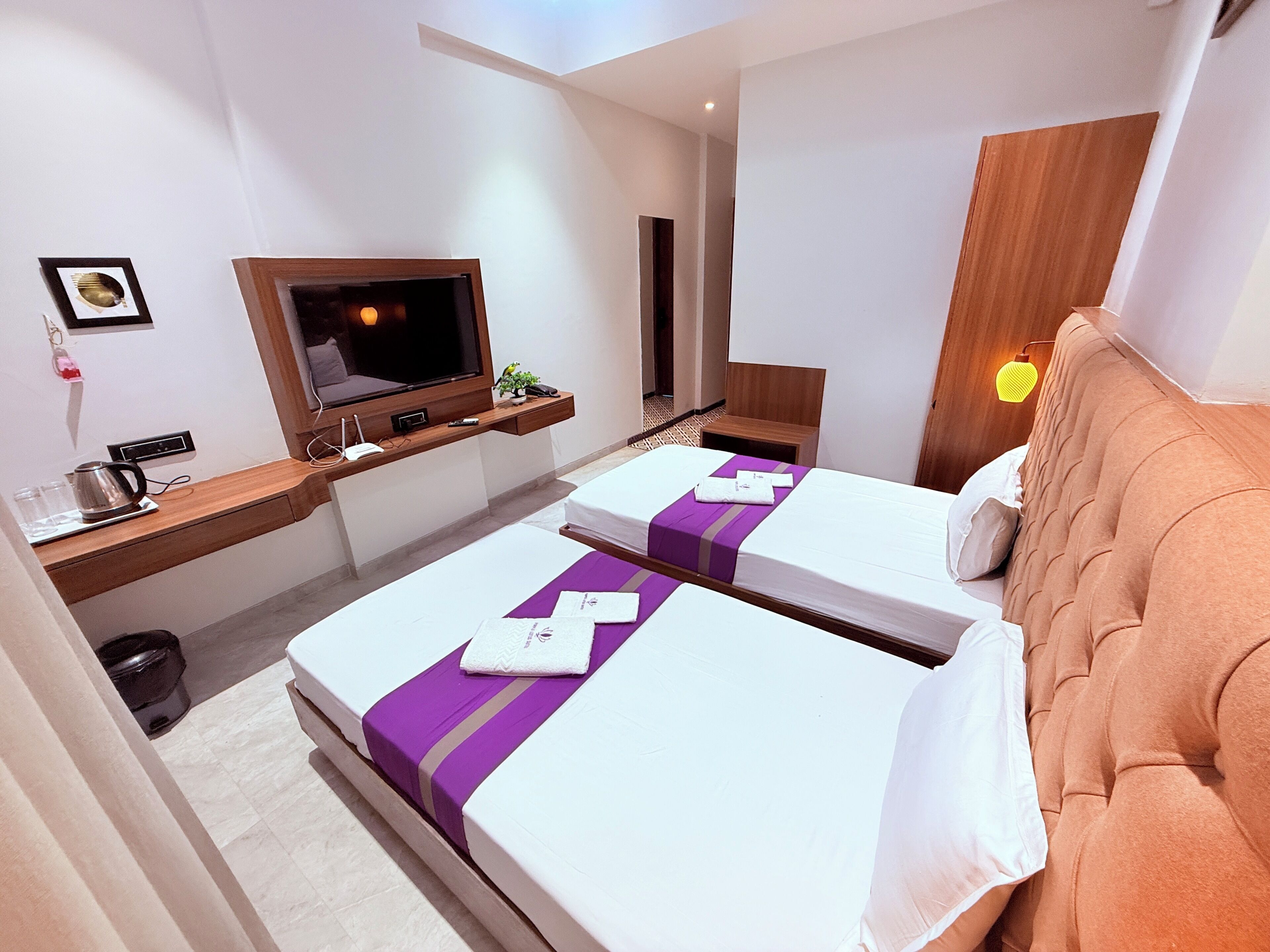 A hotel room with two beds, a TV, a desk with a kettle, and a wooden headboard.