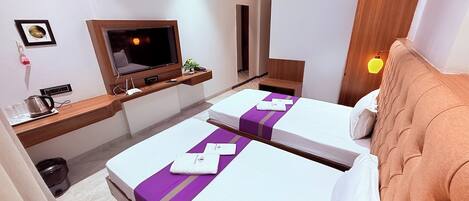 Quarto de hotel com duas camas, televisão, secretária com chaleira e cabeceira de madeira.