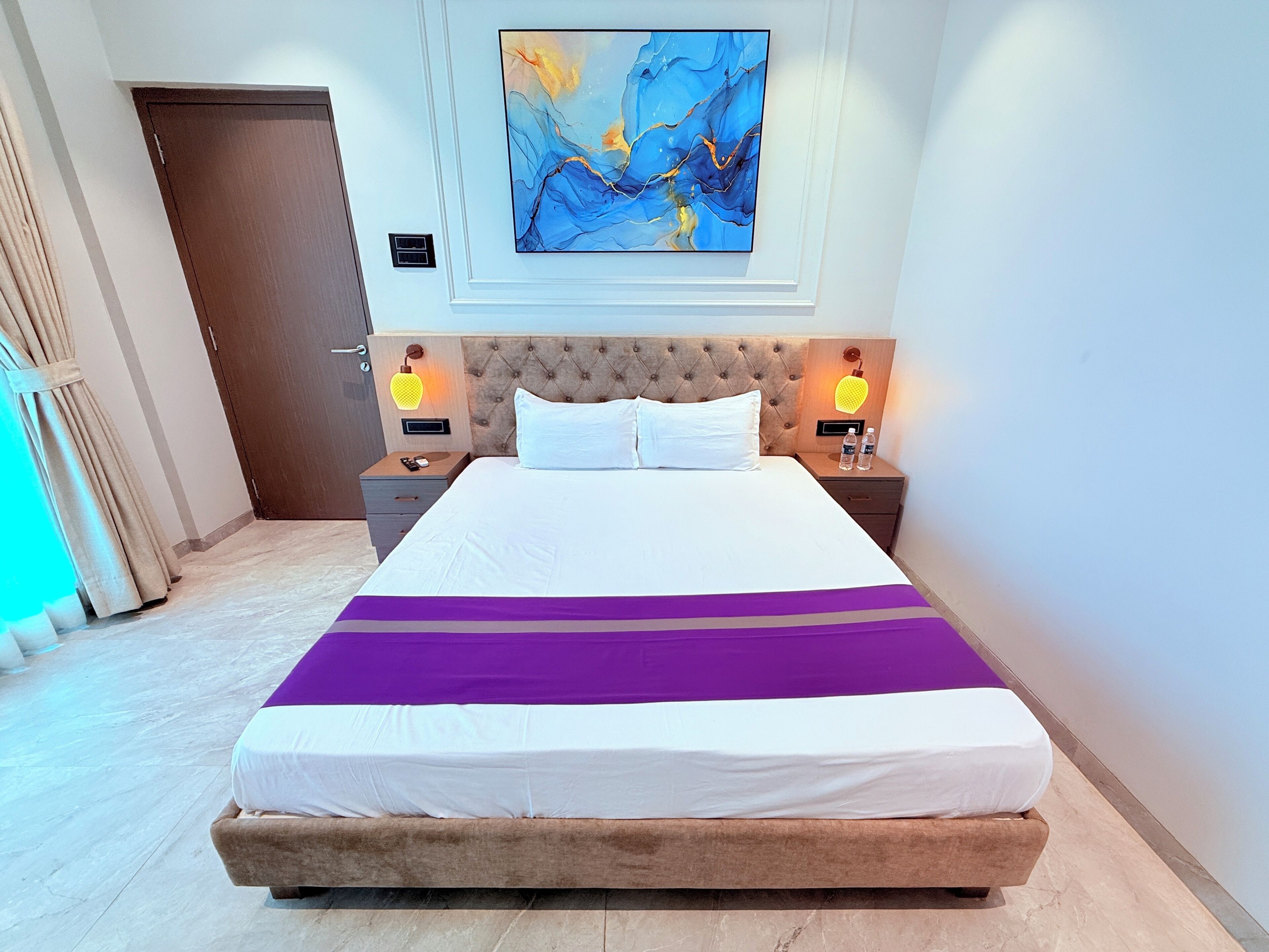 Habitación de hotel con cama, dos mesitas de noche, cabecero, cuadro abstracto en la pared y una puerta.