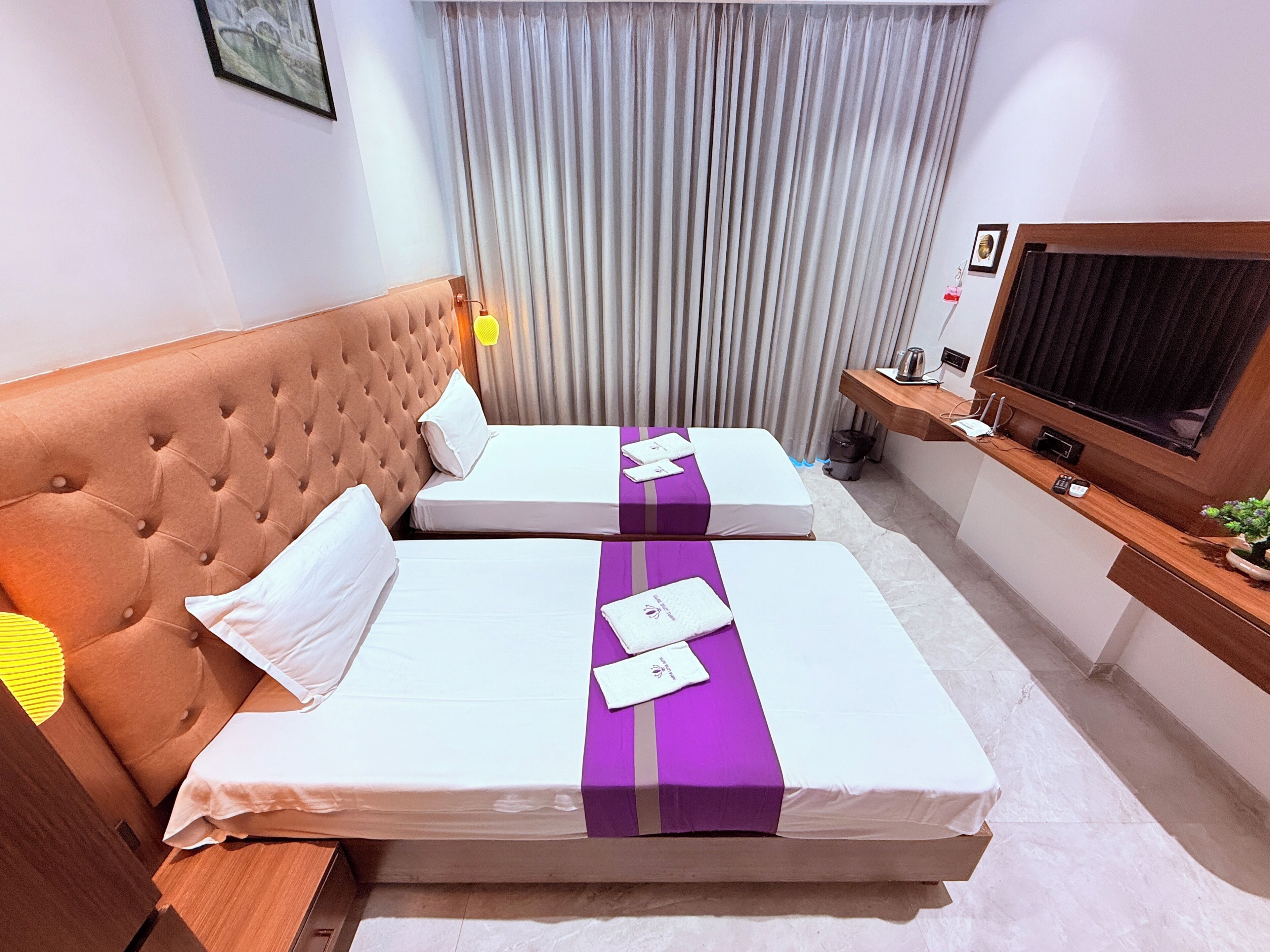 A hotel room with a tufted headboard, two beds with white and purple bedding, a television on a wooden stand, and a window with curtains.