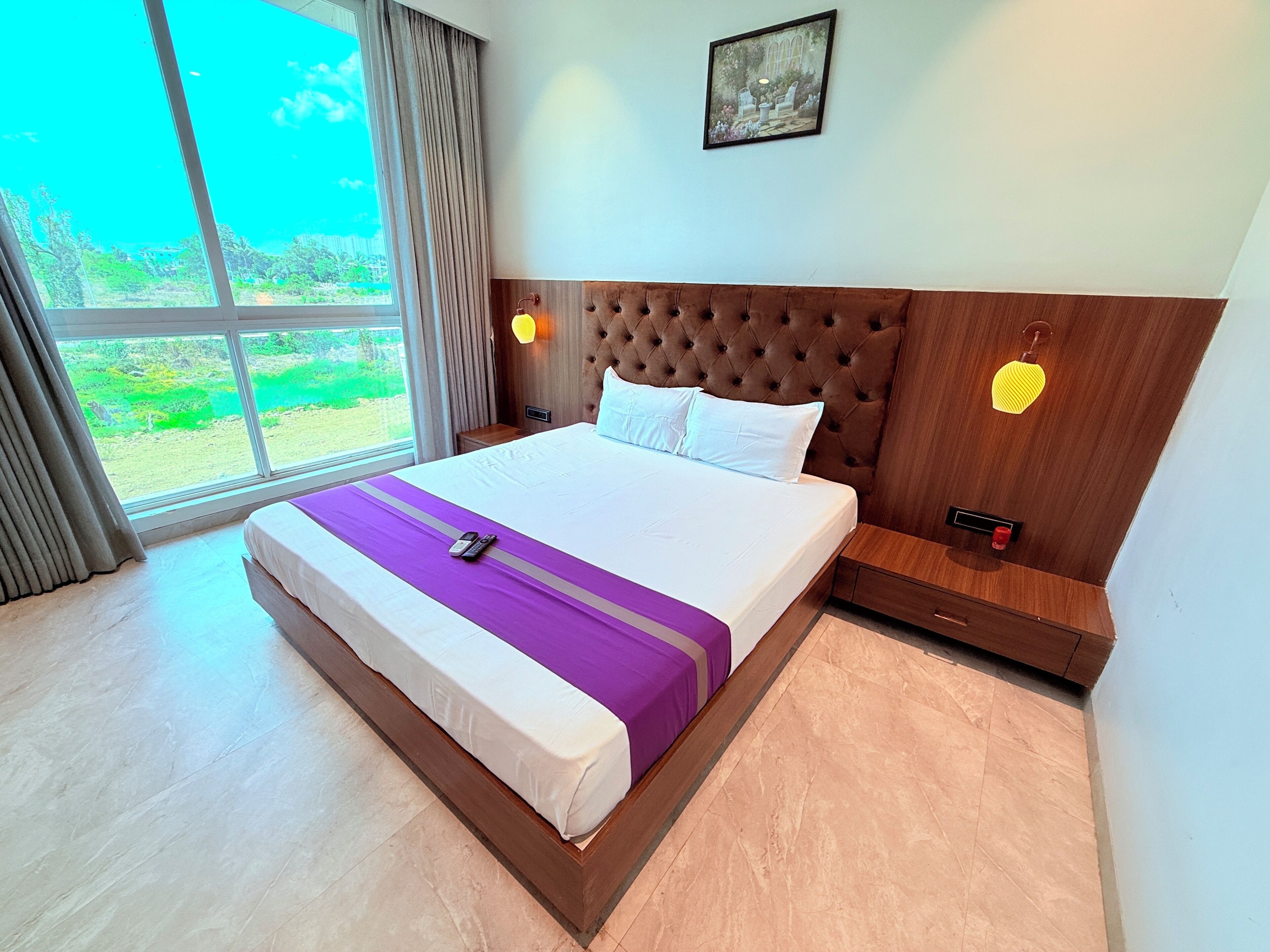 Habitación de hotel con una cama grande, vistas a zonas verdes, cabecero de madera y mesita de noche con lámpara.