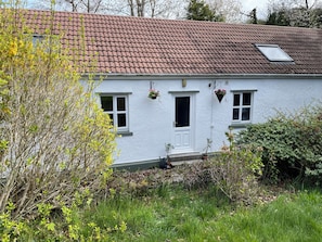 A white house with a red-tiled roof, a white door, and two windows. There are hanging flower baskets on the wall. - Inglewood Bach (Conwy)