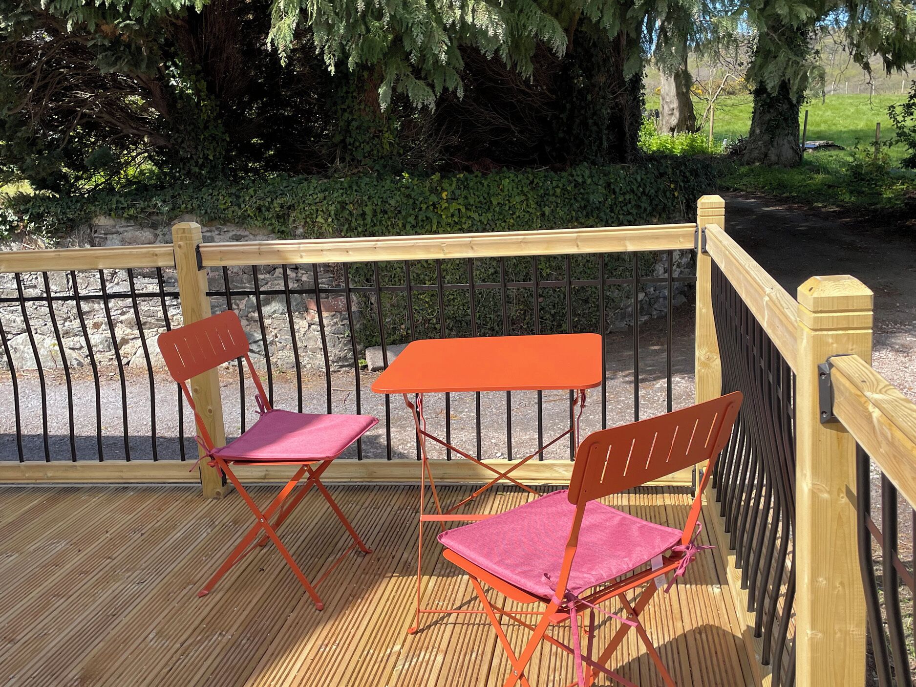 Una terraza de madera con dos sillas plegables color naranja y una mesa, rodeada por una valla metálica y follaje.