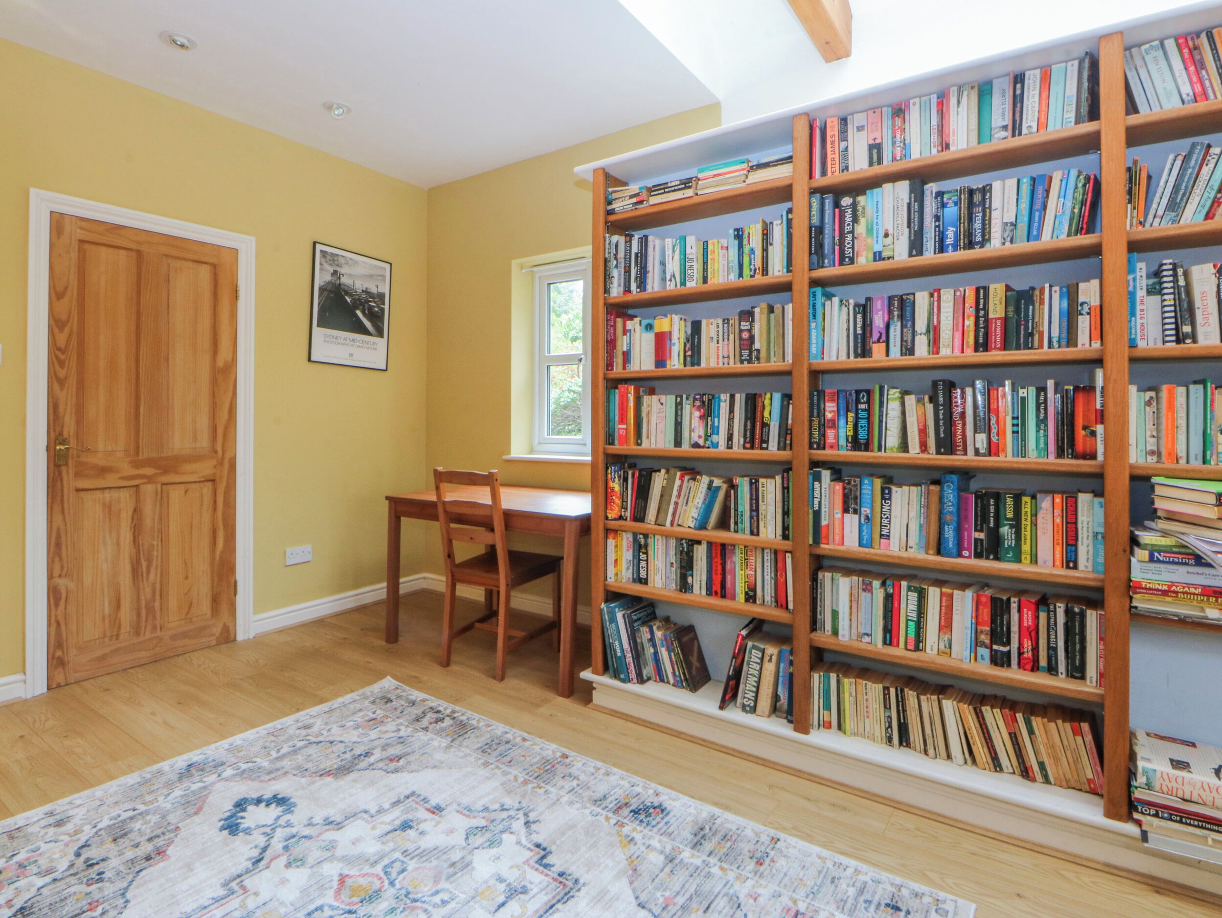 A room with a wooden bookshelf filled with books, a wooden desk, a chair, a wooden door, and a window.