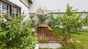 A wooden deck with a table and an umbrella, surrounded by greenery and a building.