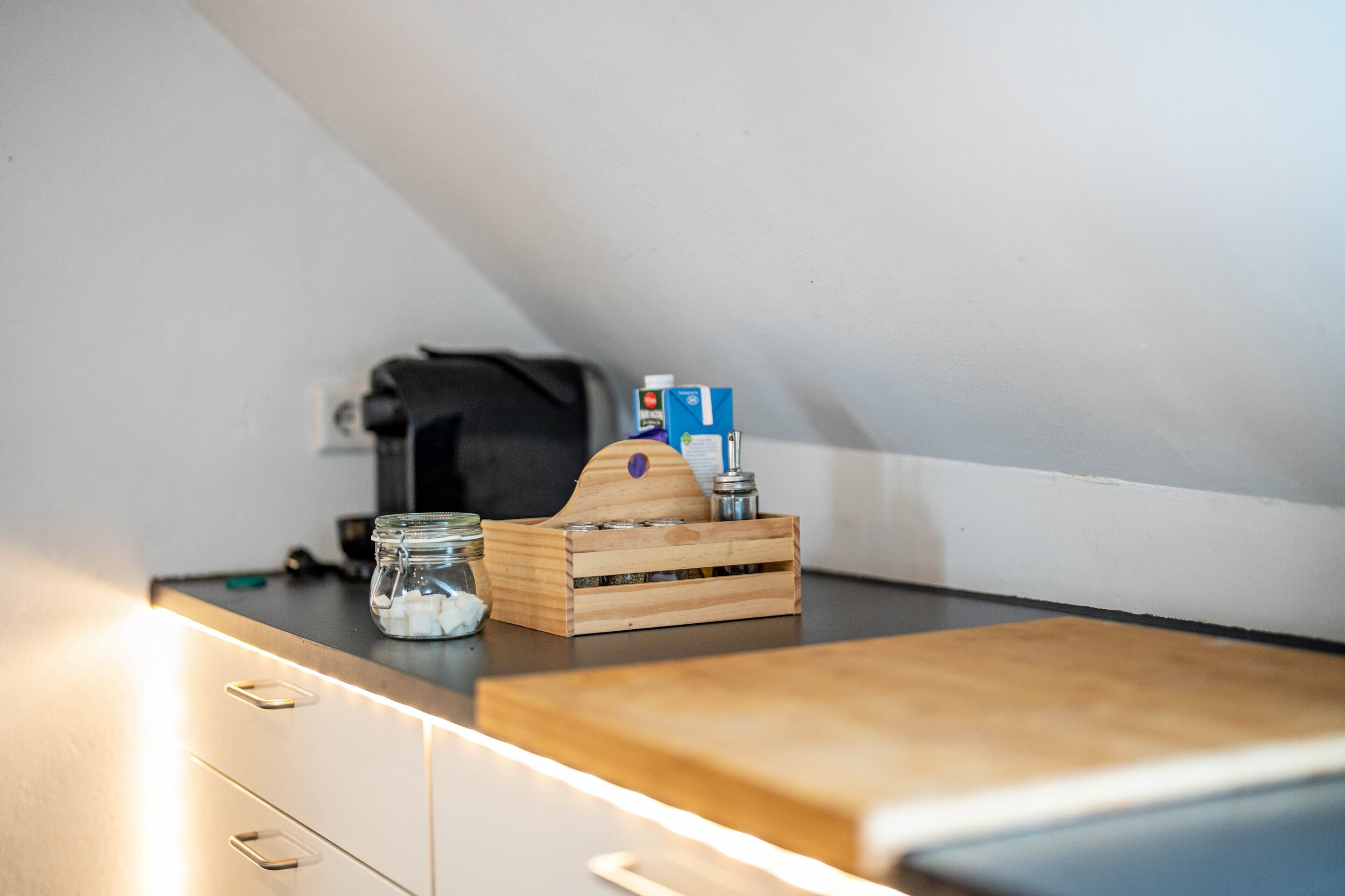 Una kitchenette con una tabla de cortar de madera, un tarro de vidrio y una caja de madera con una botella.
