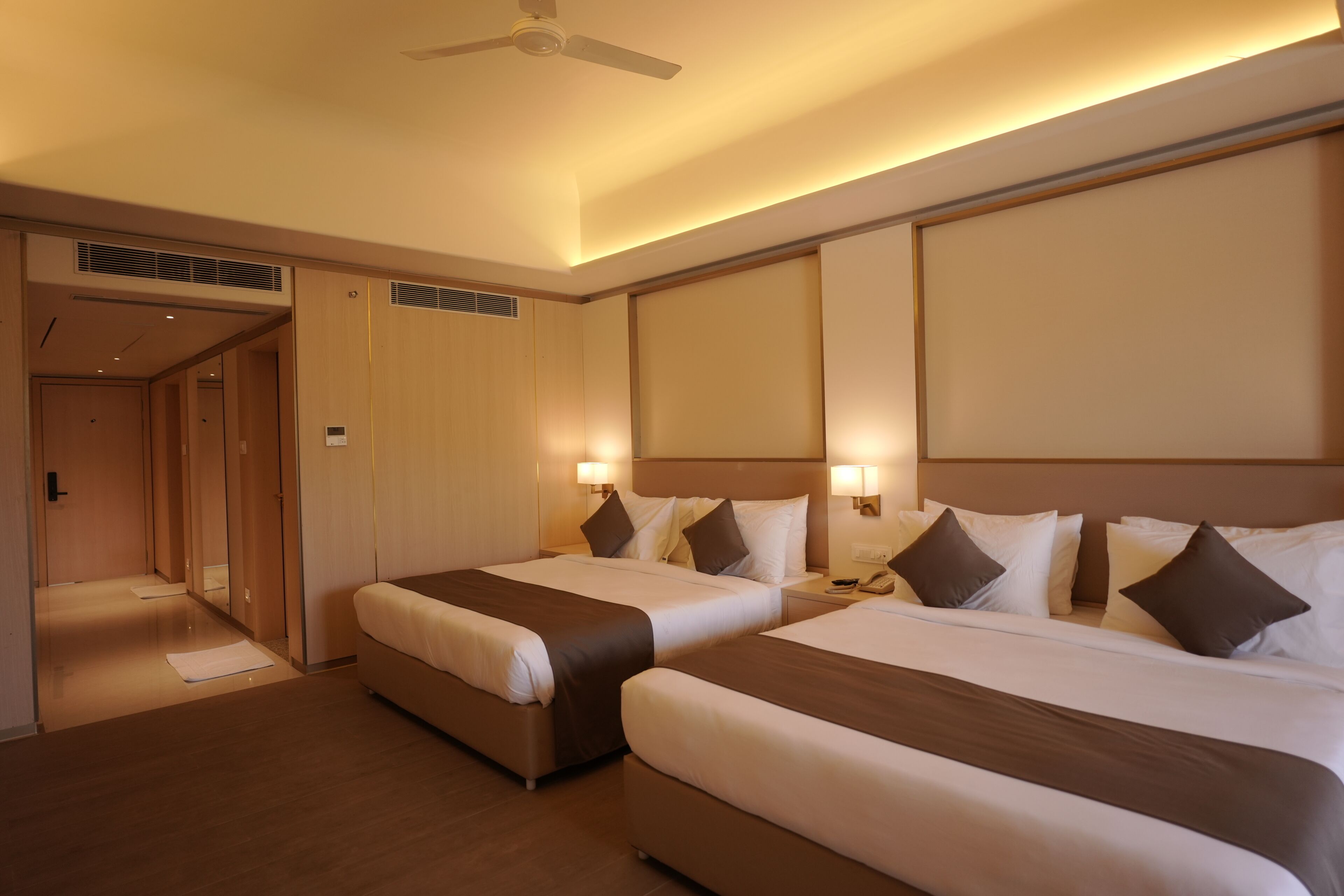 A hotel room with two beds, a ceiling fan, and a hallway leading to another room.