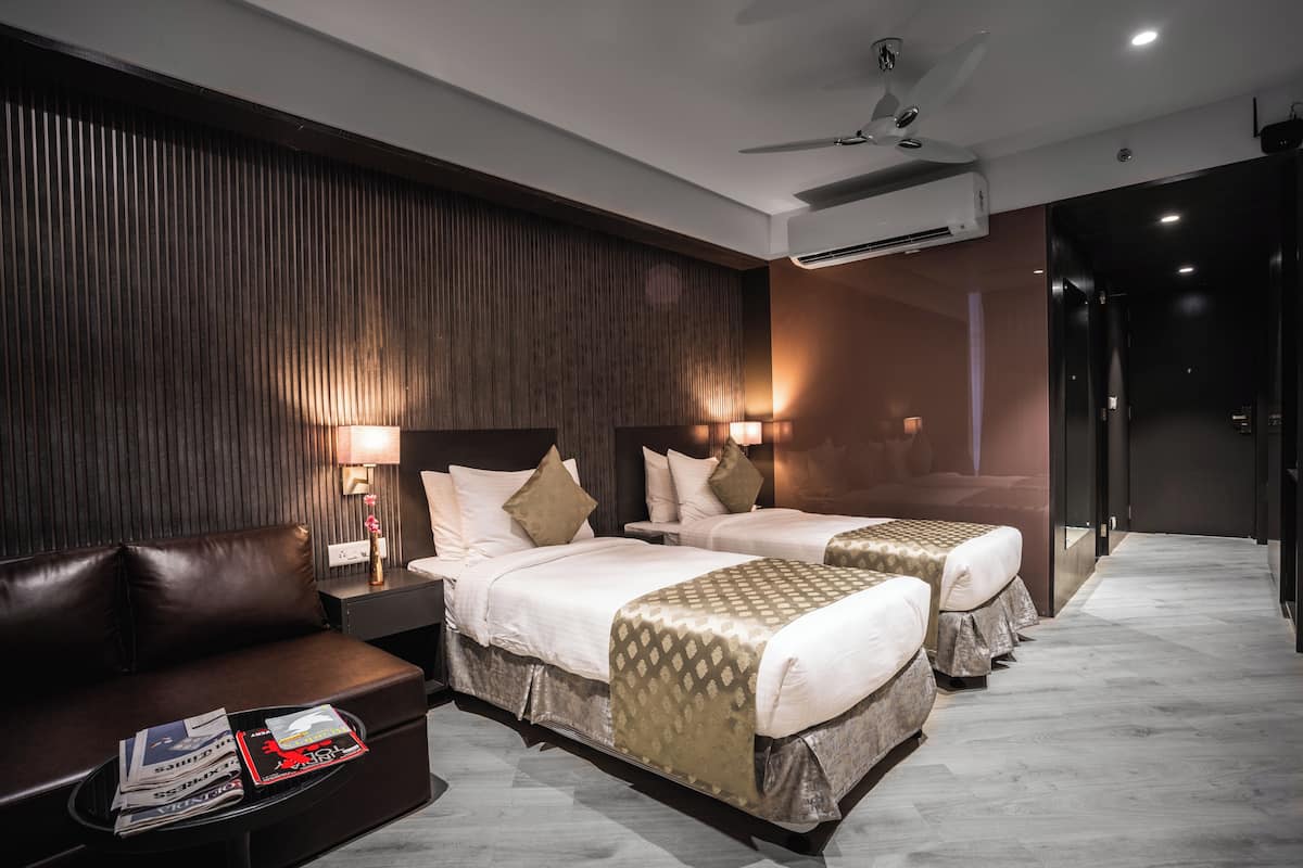 A hotel room with a sofa, two beds, bedside lamps, an air conditioner, and a ceiling fan.