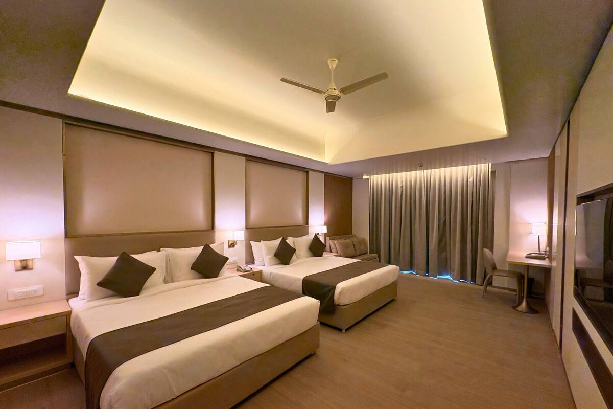A hotel room with two beds, a ceiling fan, a desk with a chair, and a large window with curtains.