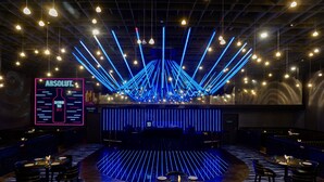 Eine Bühne mit blauem Lichtdesign, Tische mit Stühlen und ein Neon-Schild auf der linken Seite.