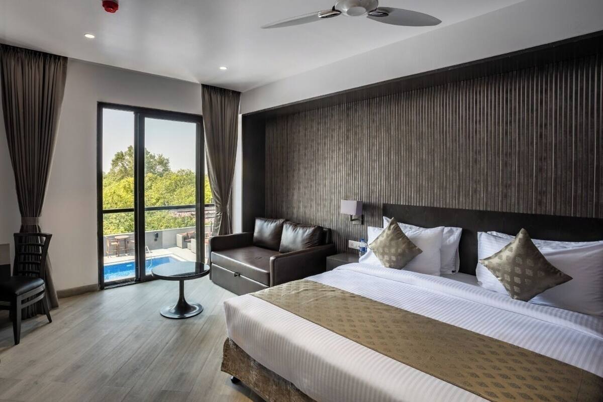 A hotel room with a bed, a sofa, a small table, a ceiling fan, and a view of the outdoors.