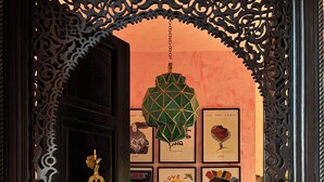 Uma porta ornamentada conduz a um quarto com tapete estampado, luminária pendente geométrica e parede decorada com obras de arte emolduradas.