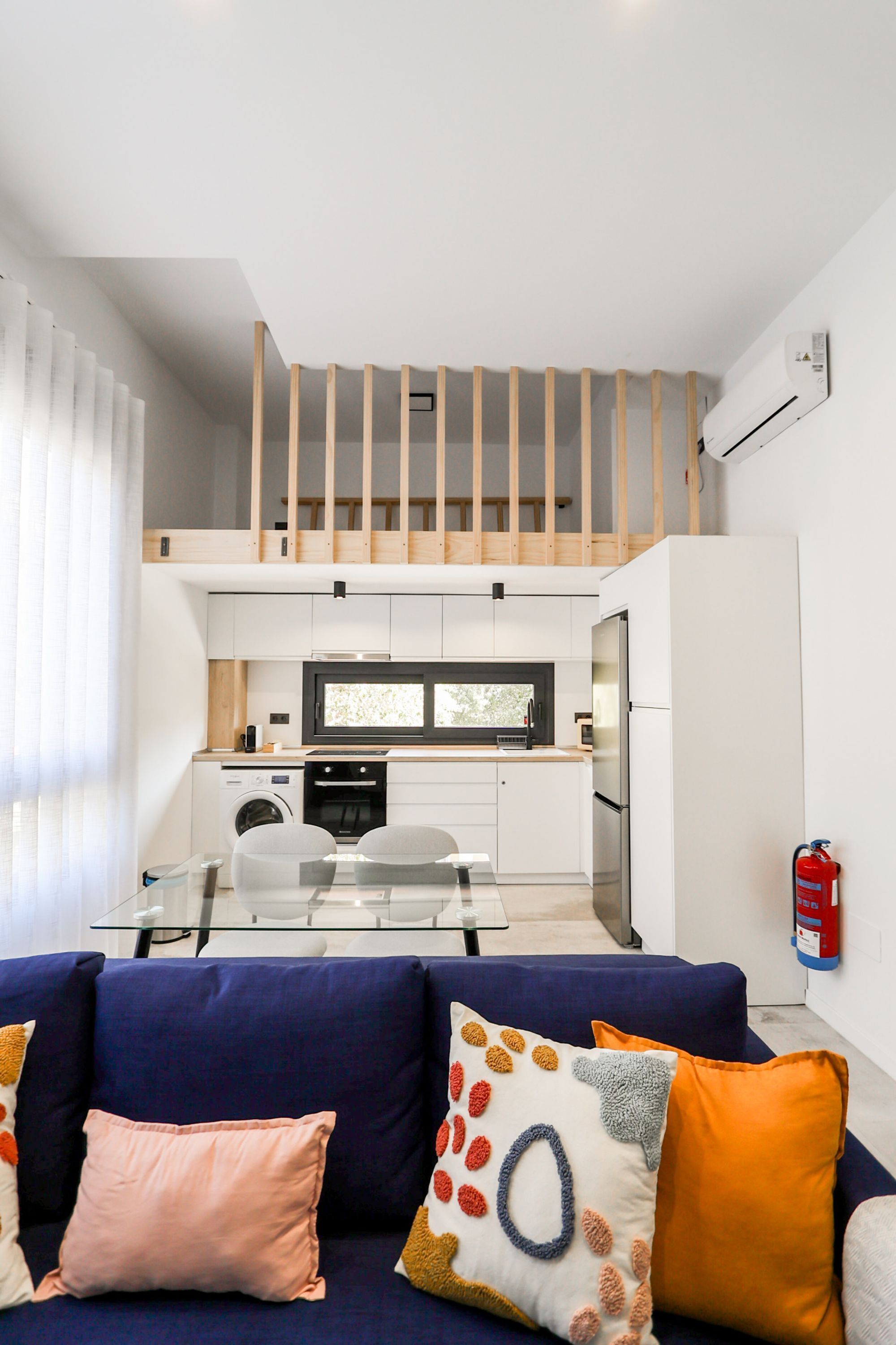 Un espace de vie compact comprenant un coin cuisine, un canapé agrémenté de coussins colorés et un escalier menant à un niveau supérieur.