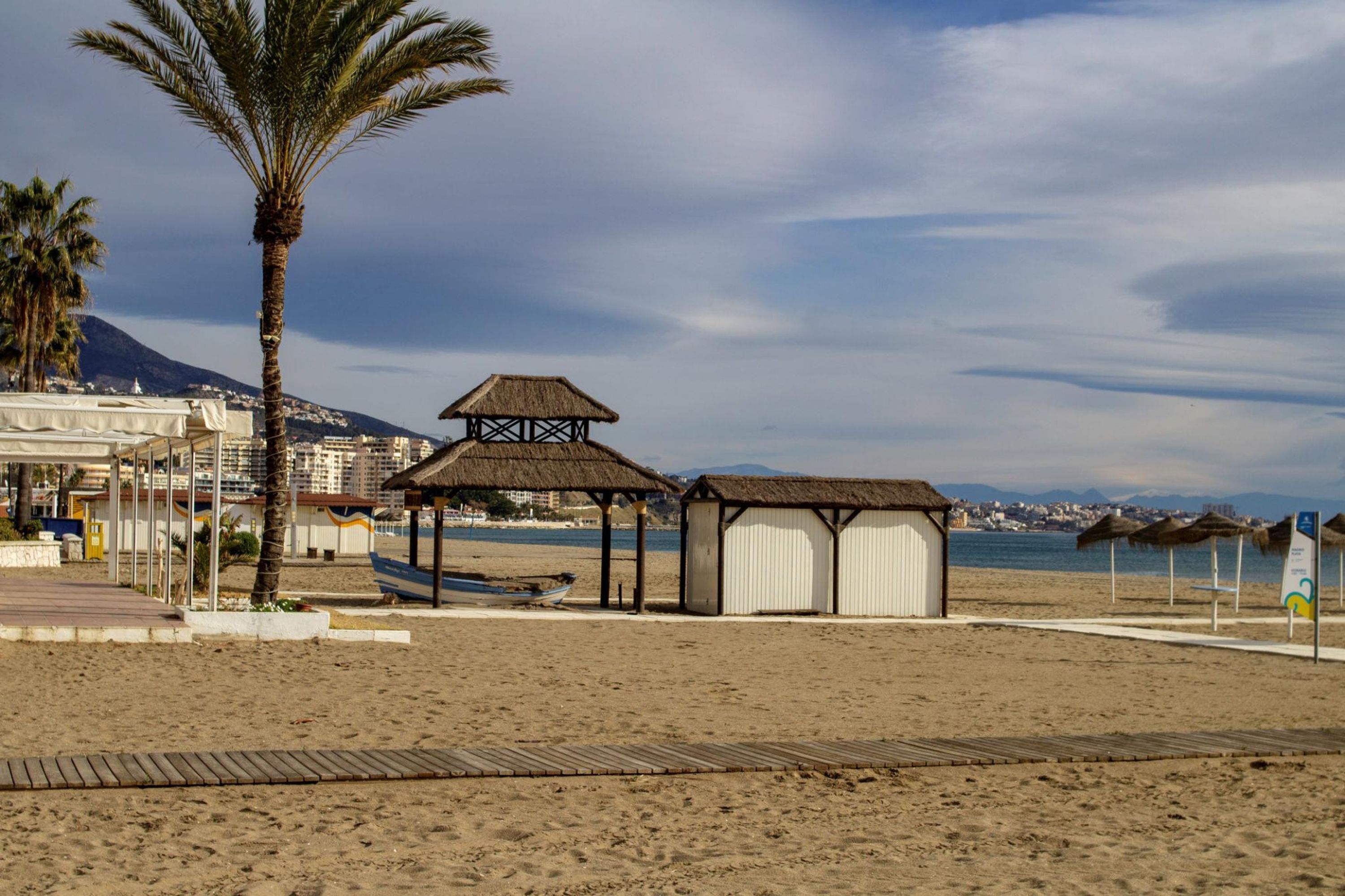 A beach with a wooden walkway, palm trees, a thatched-roof shelter, and a small building.