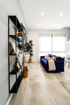 Ein modernes Wohnzimmer mit einem blauen Sofa, einer schwarzen Regaleinheit und einem großen Fenster mit durchsichtigen Vorhängen.