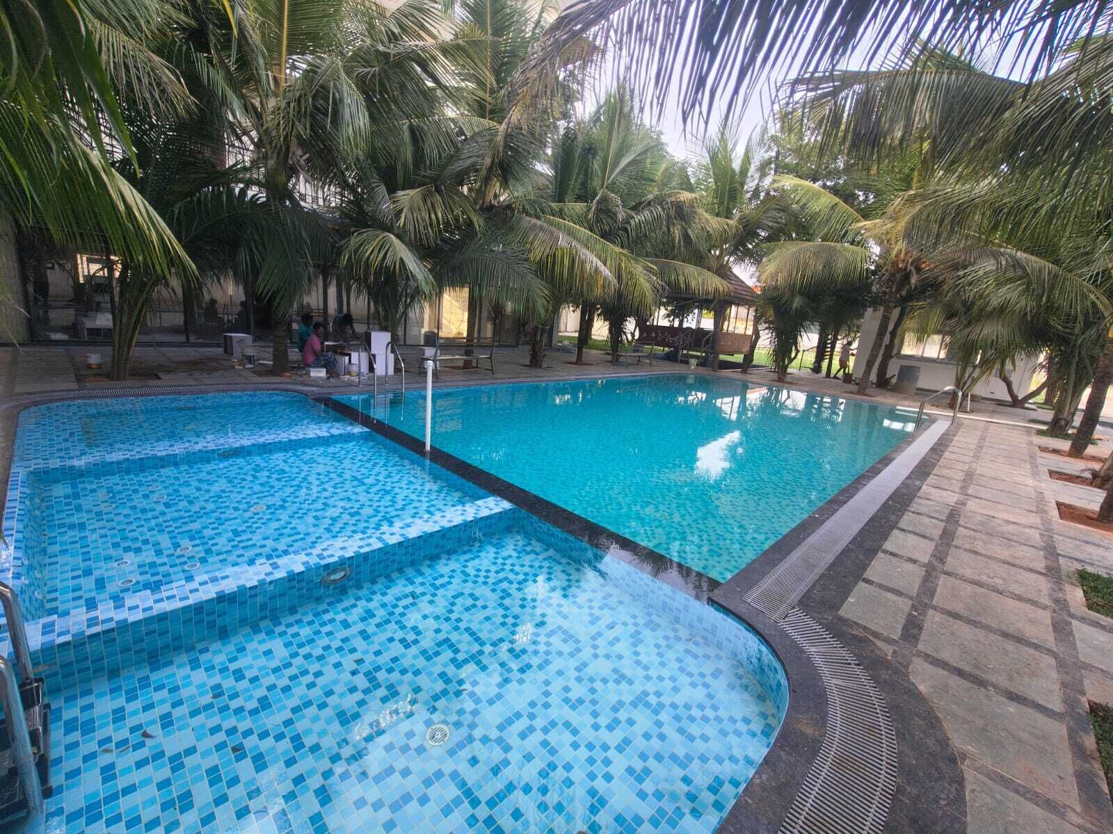 An indoor swimming pool with tiled flooring, surrounded by palm trees and a covered seating area.