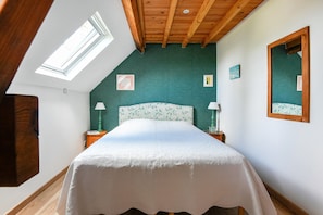 A bedroom with a sloped ceiling, a skylight, a bed with a floral headboard, two bedside tables with lamps, and framed pictures on the walls.