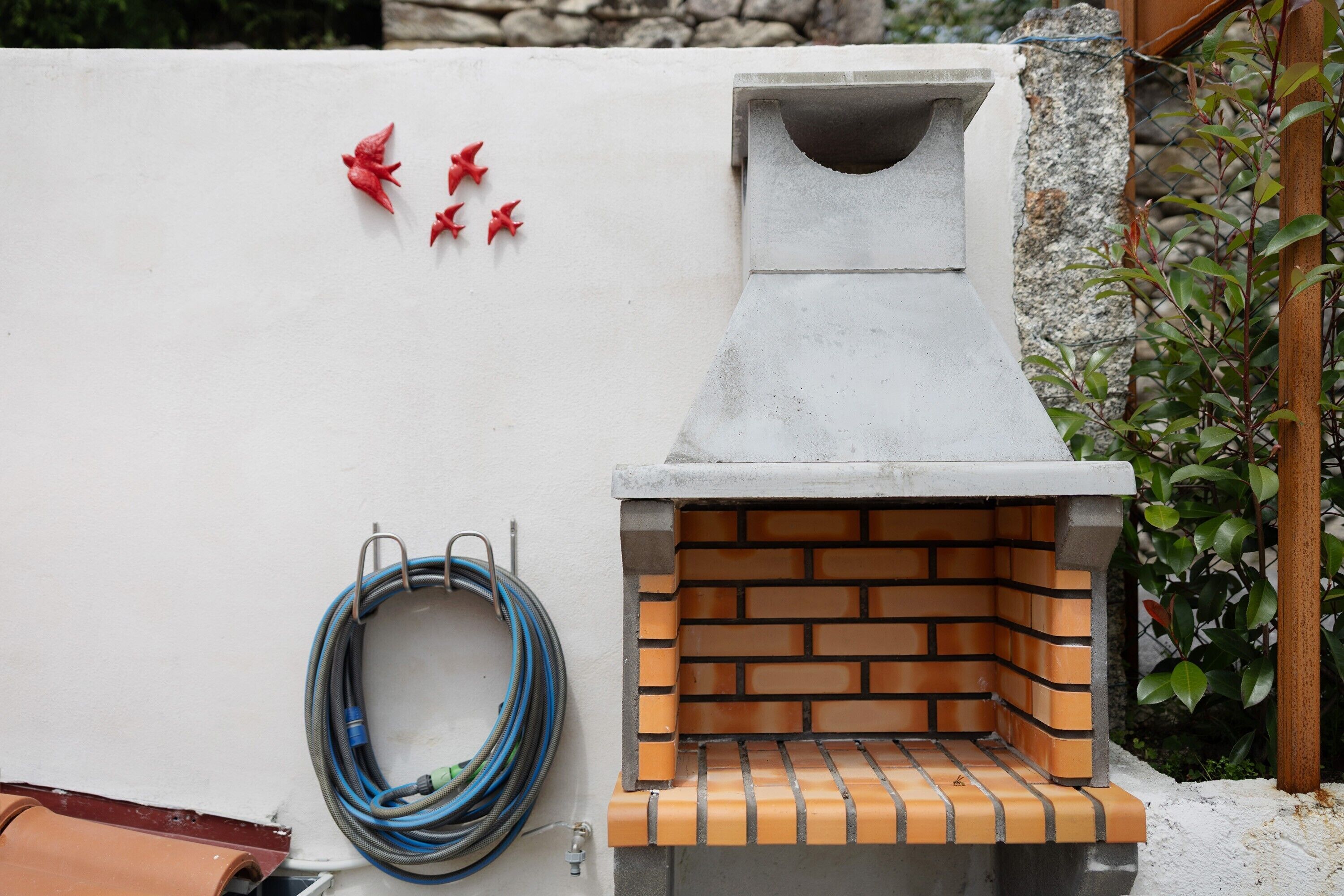 A brick-built outdoor grill with a chimney, a coiled garden hose, and red decorative elements on a white wall.