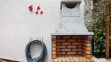 A brick-built outdoor grill with a chimney, a coiled garden hose, and red decorative elements on a white wall.