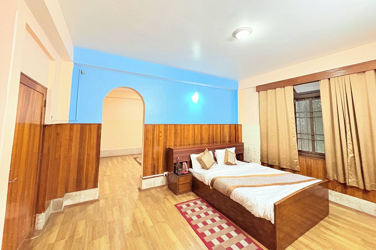 A bedroom with a wooden bed, a nightstand, a red and white patterned rug, a window with curtains, and a blue wall with an archway.