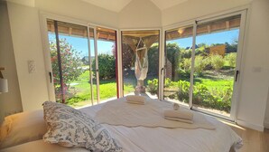 Une chambre avec un grand lit, un fauteuil suspendu et une vue sur un jardin, accessible par des portes-fenêtres coulissantes.