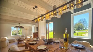Un coin repas avec une table en bois dressée pour six personnes, une bouteille de vin et une vue sur un paysage verdoyant par de grandes fenêtres.
