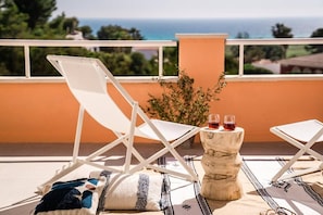 A balcony with two white lounge chairs, a small round table with two glasses of red wine, and a view of the sea and greenery. - Refugi Blau, sea view in Son Bou (Son Bou)
