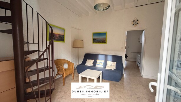 A living room with a blue sofa, a wicker chair, a small white table, and a staircase. There are two framed pictures on the wall. - Apartment in Soulac town center, all on foot (Soulac-sur-Mer)