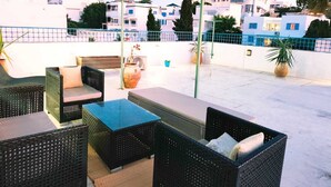 En udendørs terrasse med fletmøbler, et bord med glasplade og plantekrukker.