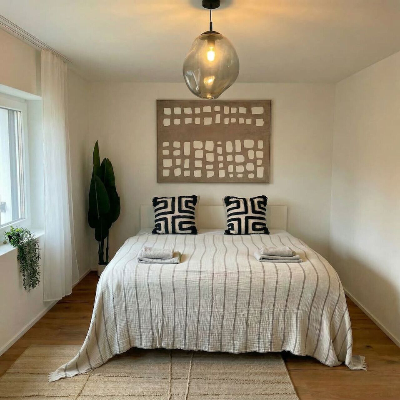 Une chambre à coucher avec un lit, un luminaire suspendu, une œuvre d’art murale et une fenêtre avec des rideaux.