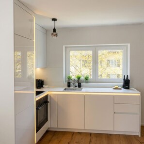 A modern kitchen with white cabinetry, a built-in oven, a double sink, and a window with a view of trees. - Modern 2BR | Balcony - Quiet - 7min ZRH Airport (Opfikon)
