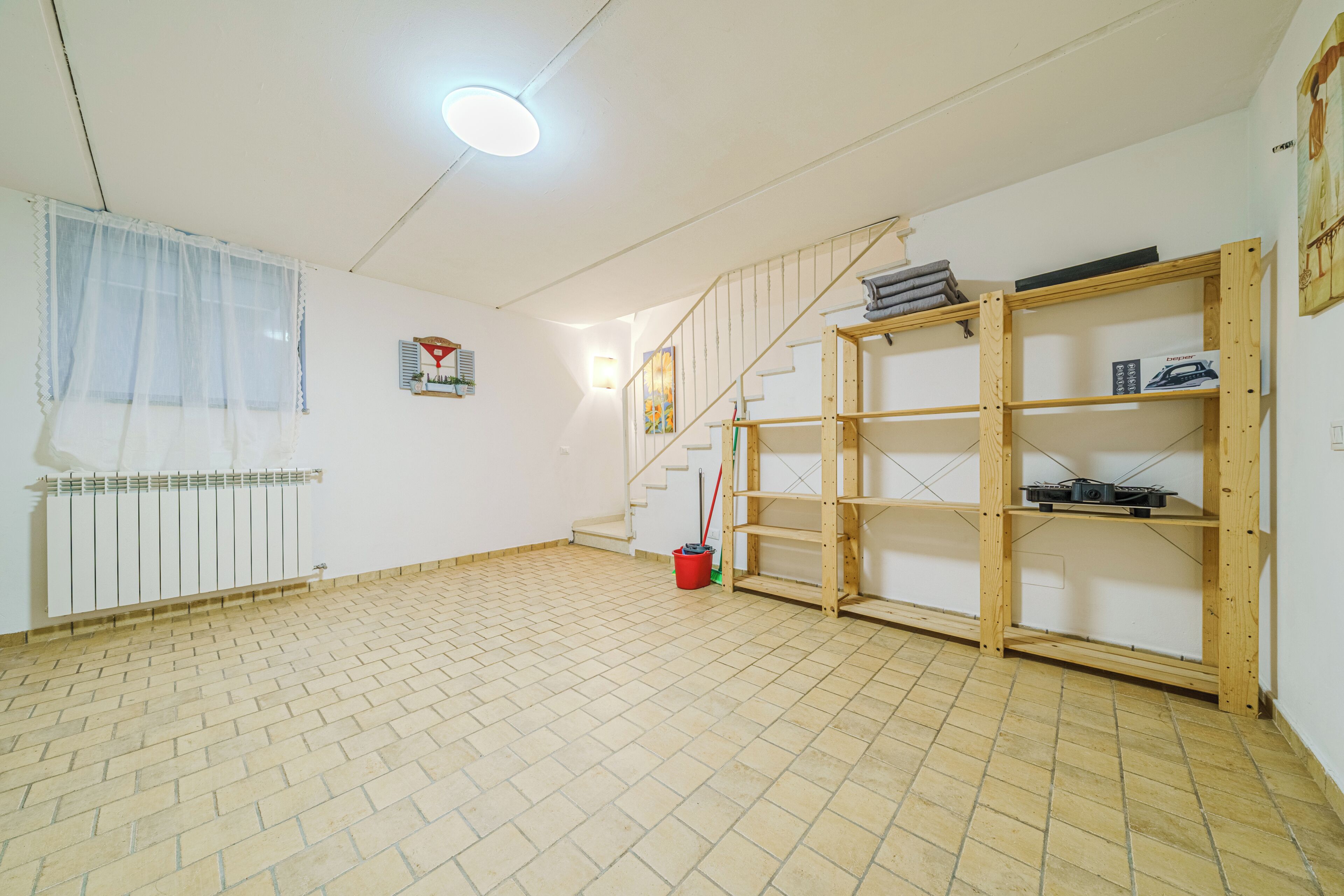 Una habitación con suelo de baldosas, una escalera, un radiador, una ventana con cortina ligera y un estante de madera.
