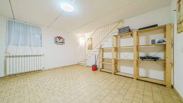 Una habitación con suelo de baldosas, una escalera, un radiador, una ventana con cortina ligera y un estante de madera.
