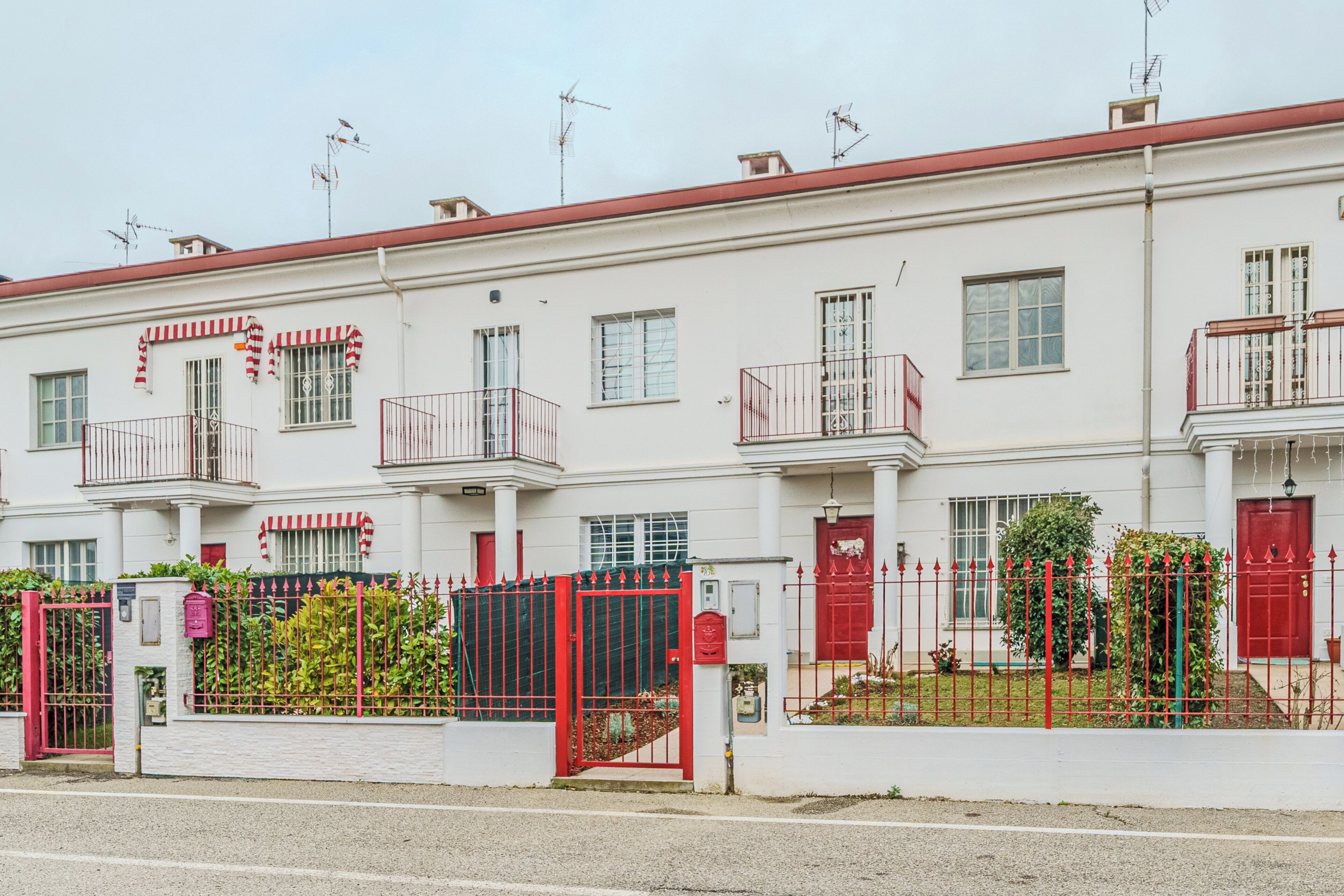 Una hilera de edificios blancos con balcones, un portón rojo y un pequeño jardín.