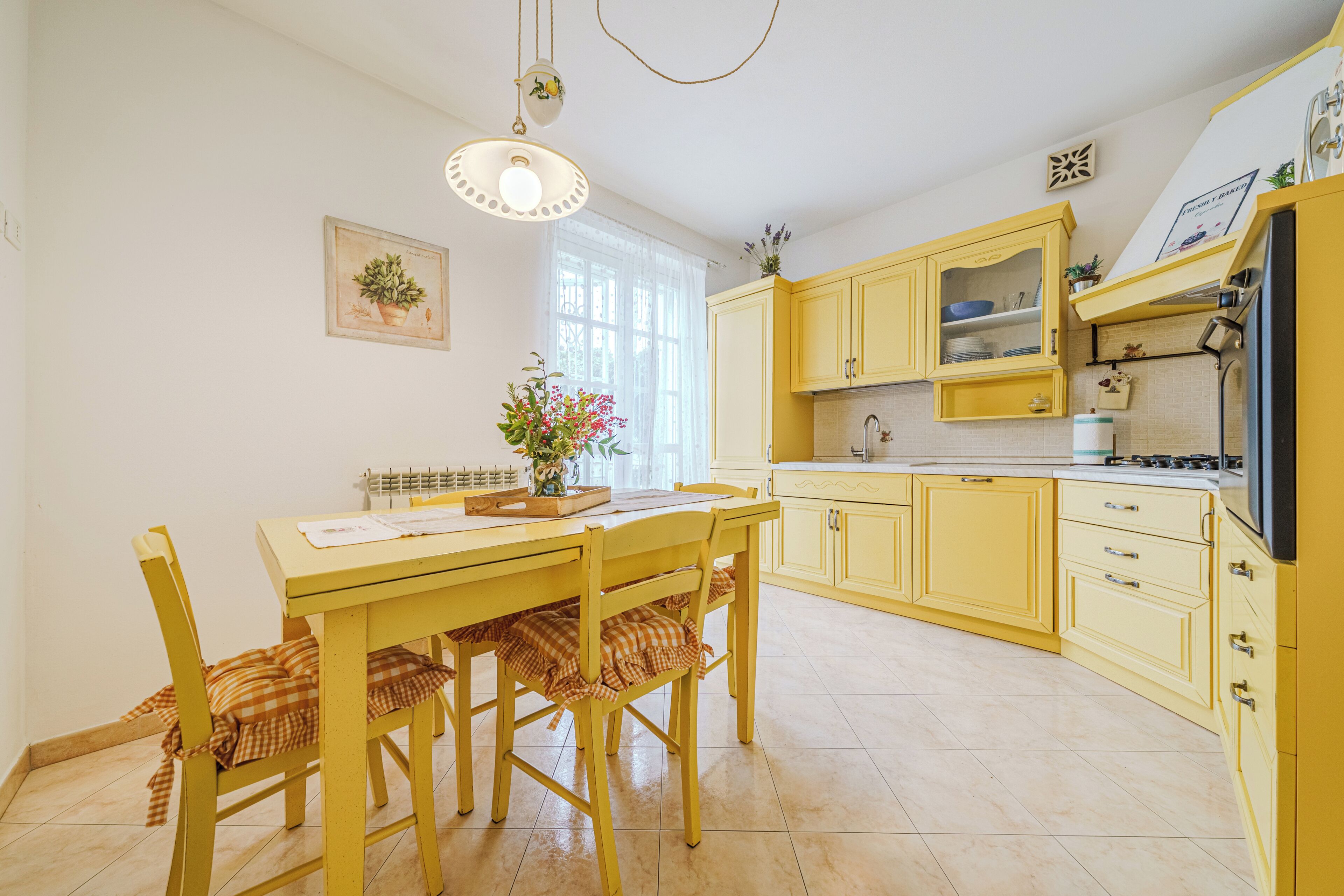 A kitchen with yellow cabinets, a dining table with chairs, a hanging light fixture, and a window with curtains.