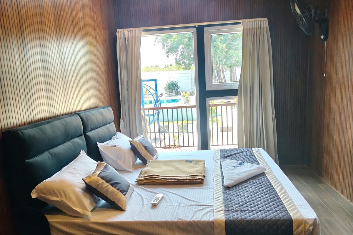 Una habitación de hotel con cama, ventana con cortinas, vistas al exterior y un ventilador en la pared.