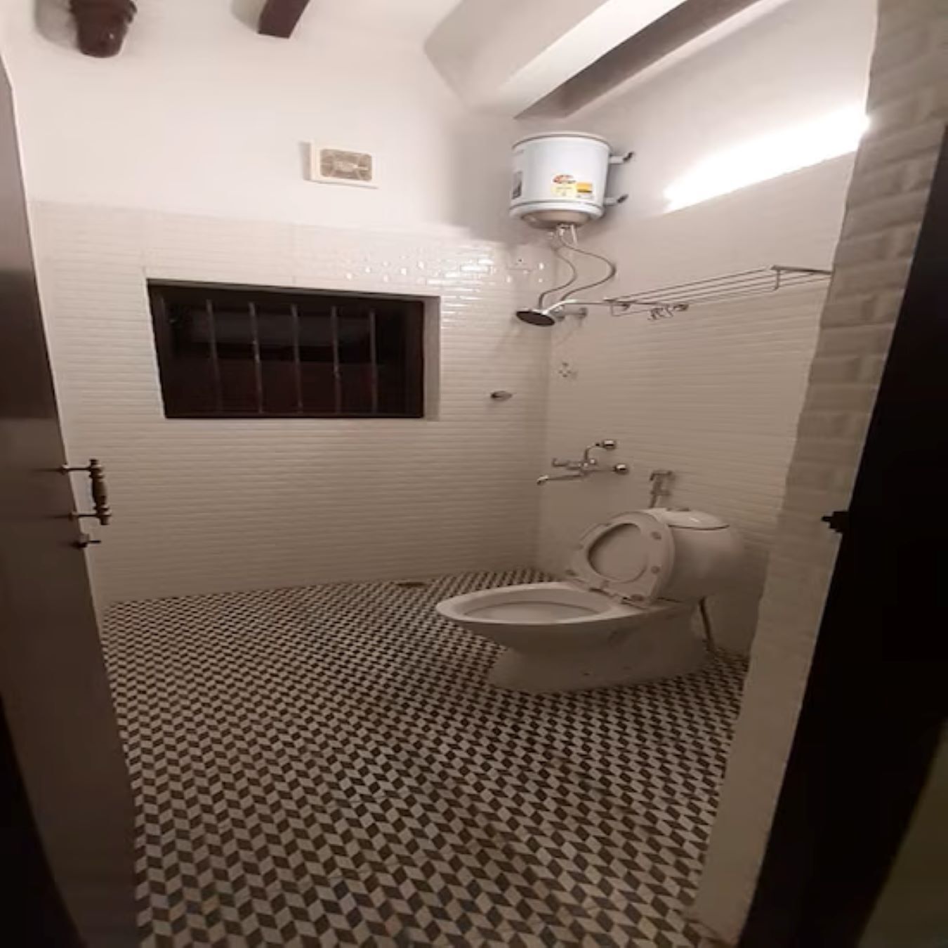 Une salle de bain avec des toilettes, une fenêtre avec des barreaux, un carrelage au sol et un chauffe-eau fixé au plafond.