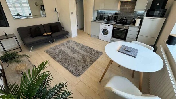 Een ruimtelijk ingerichte leefruimte met een bank, een ronde eettafel, een wasmachine en een kitchenette.