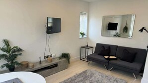 Een moderne woonkamer met een grijze bank, een flatscreen televisie, een ronde witte tafel en een ruig, grijs vloerkleed.