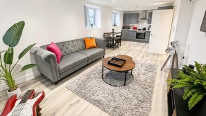 A modern living room with a grey sofa, a coffee table, and a dining area with a small table and chairs. - The Studios at Taylor’s Court (Canvey Island)