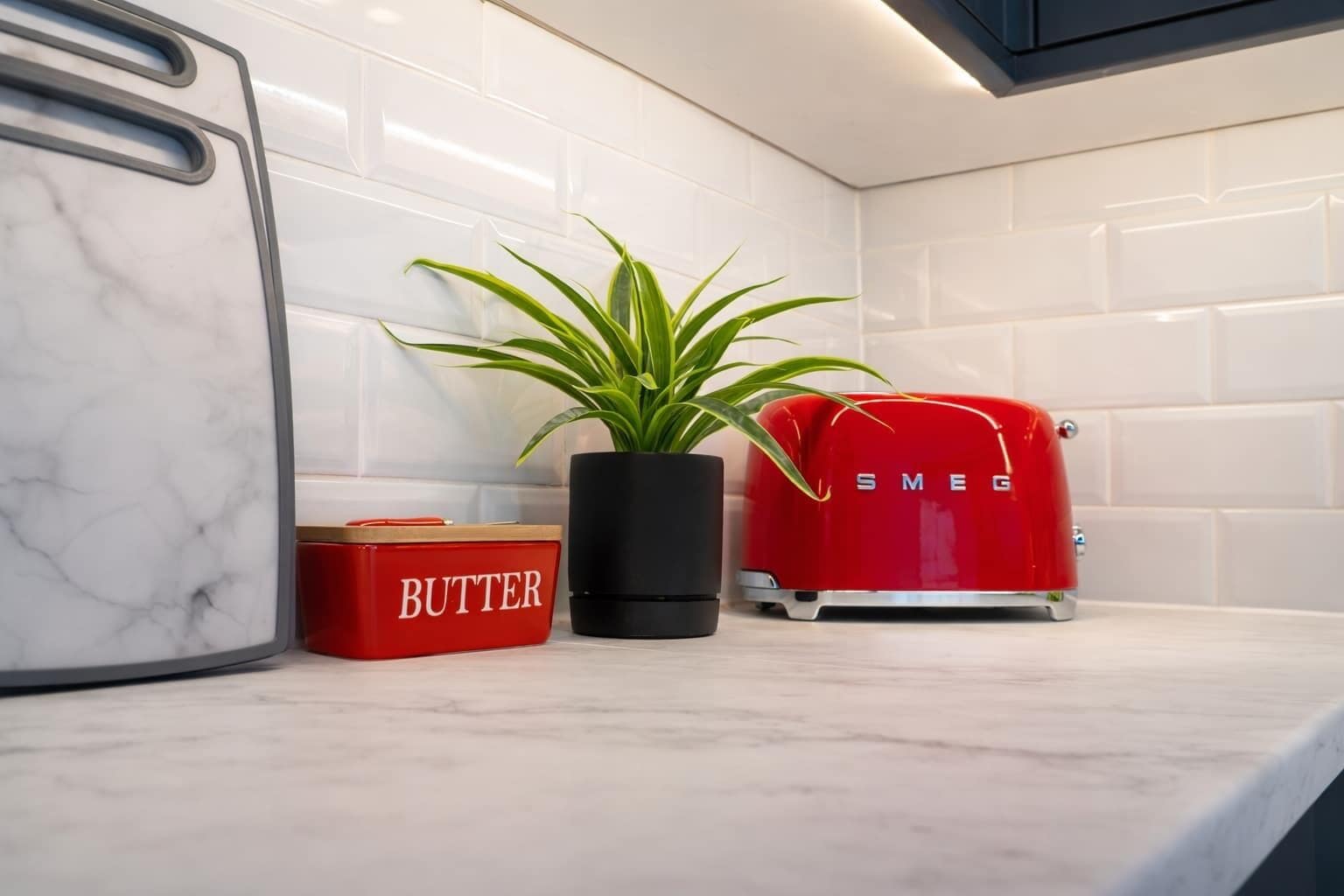 Un plan de travail de cuisine avec un pare-feu en marbre, un pot à beurre rouge, une plante en pot et un grille-pain rouge de la marque Smeg.