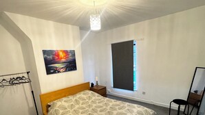 Ein Schlafzimmer mit Bett, Fenster mit Jalousie, Nachttisch, Stuhl und einer Wandlampe.