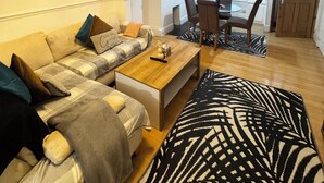 Ein Wohnzimmer mit einer Couch, einem Couchtisch und einem gemusterten Teppich. Es gibt einen Essbereich mit Stühlen und einem Tisch.