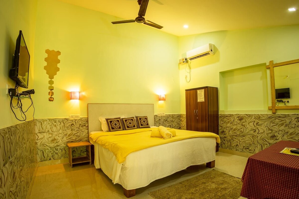 A hotel room with a bed, bedside lamps, a ceiling fan, an air conditioning unit, a wooden wardrobe, a small table with a red tablecloth, and a wall-mounted TV.