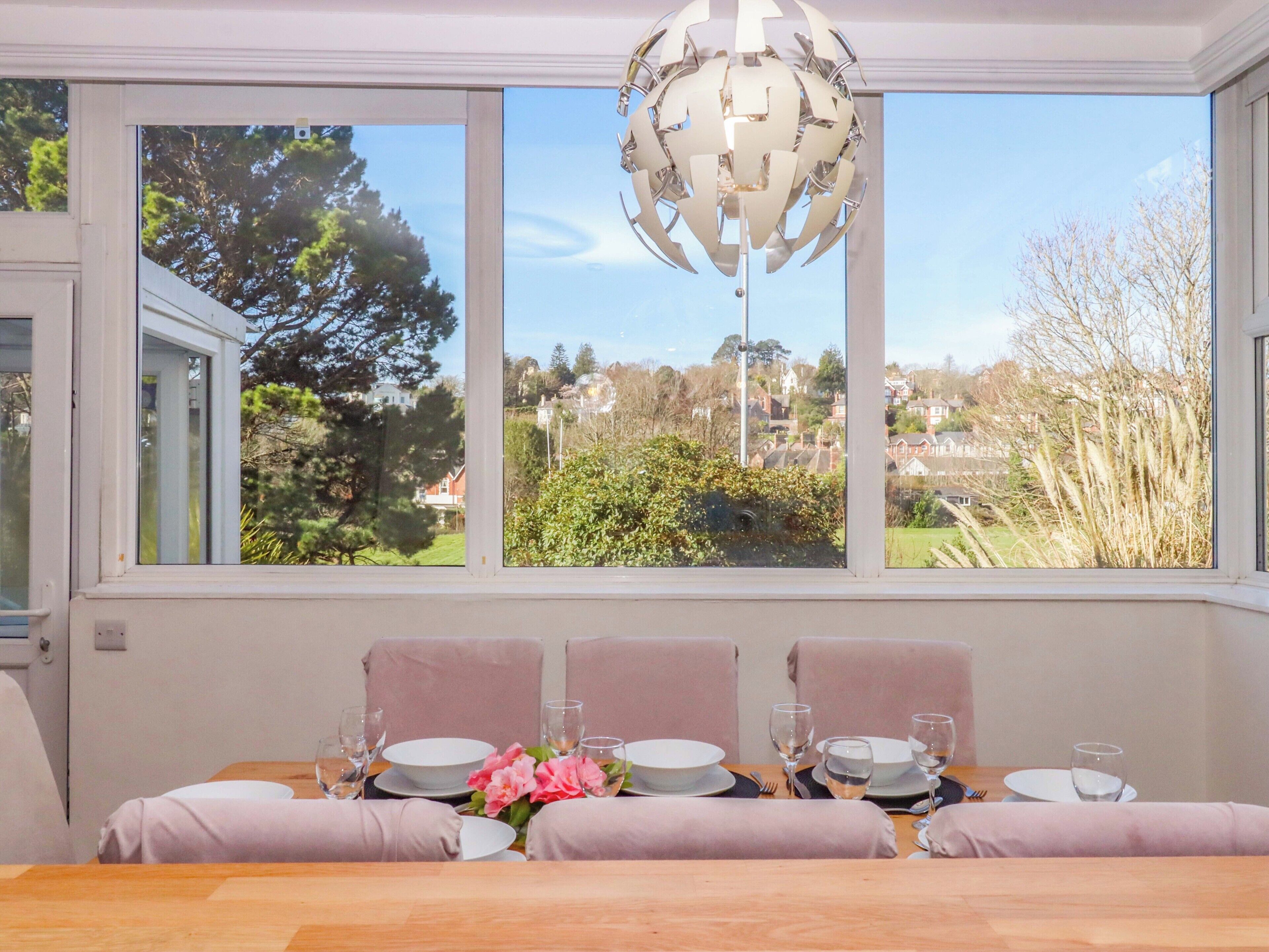 Comedor con mesa dispuesta para cuatro, una araña y vistas a un barrio residencial a través de amplias ventanas.