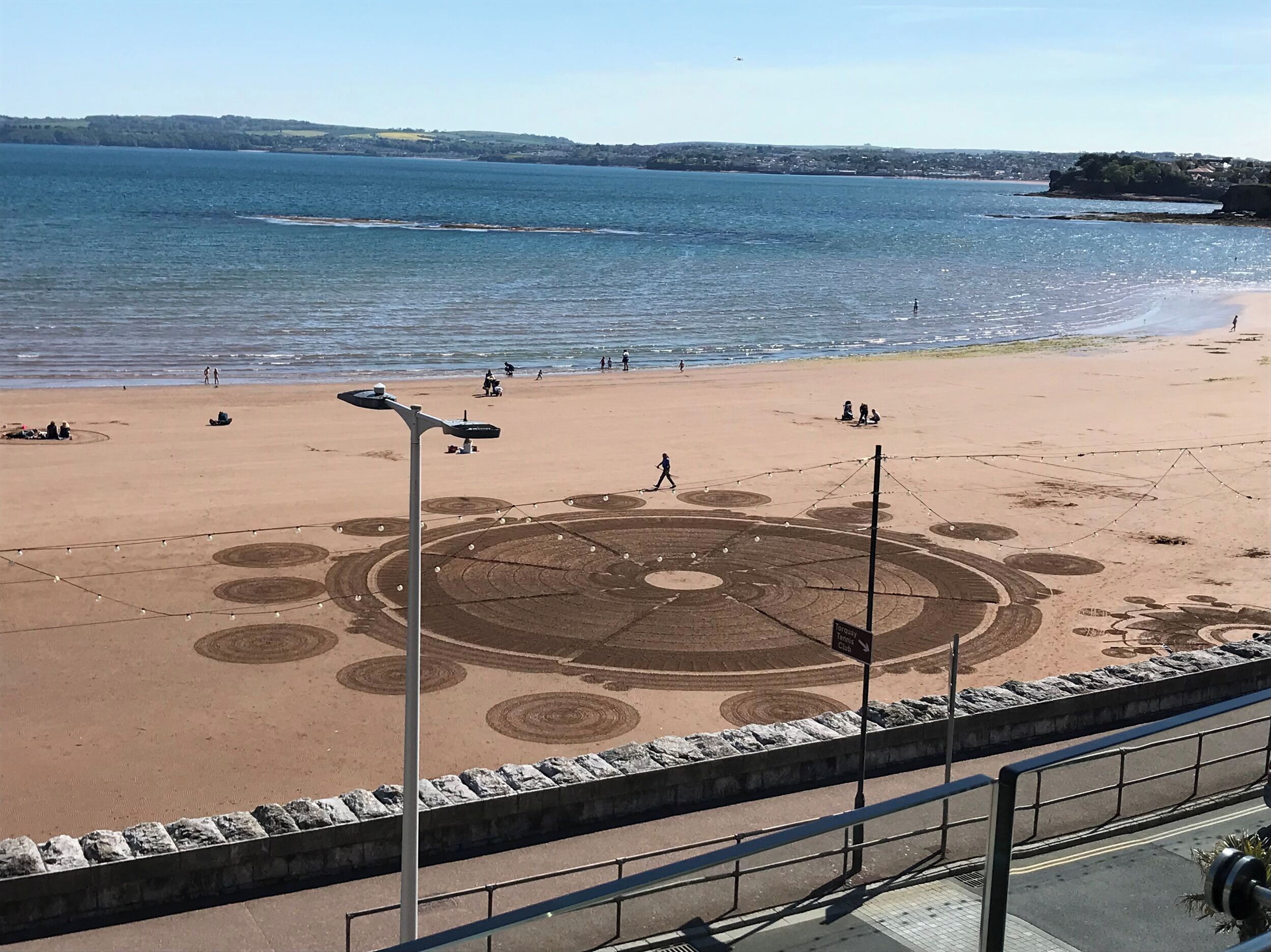 Una platja amb un dibuix circular a la sorra, un passeig pavimentat i vista al mar.